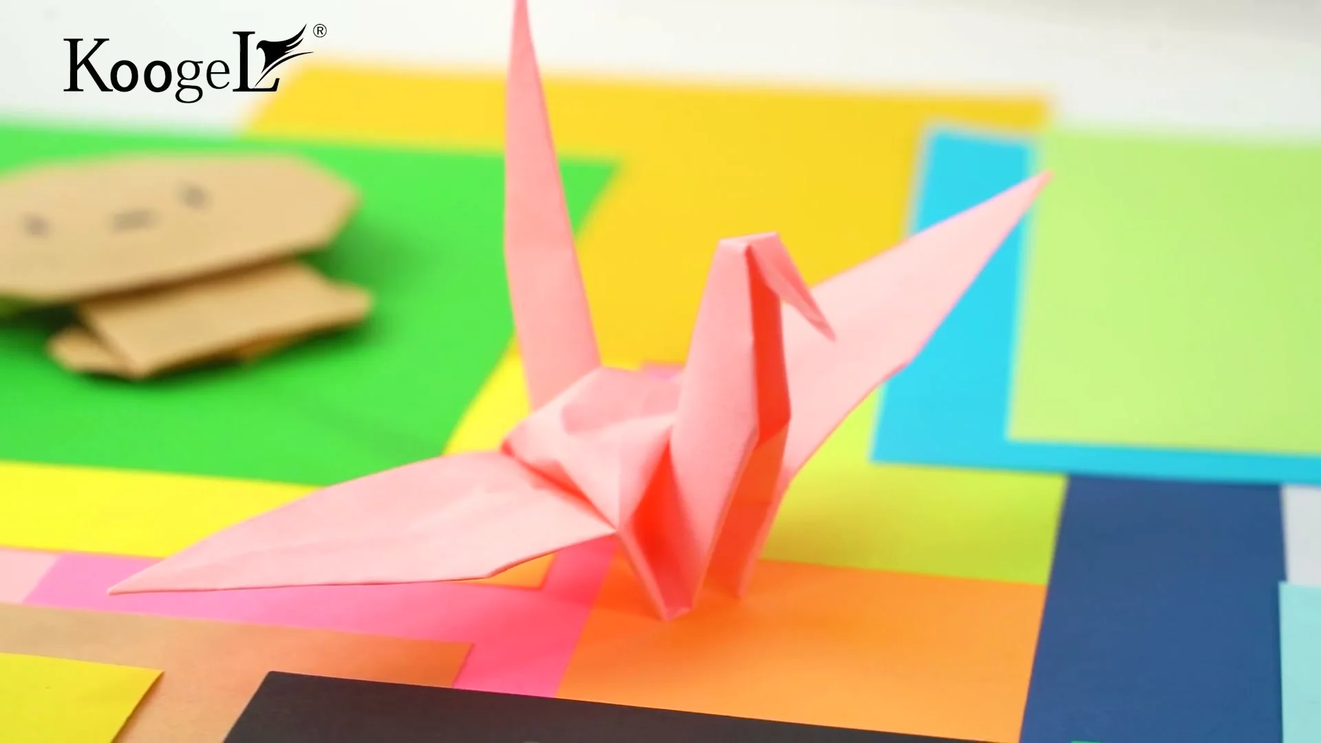 A pink origami crane folded from thin paper, resting on sheets of colored paper.